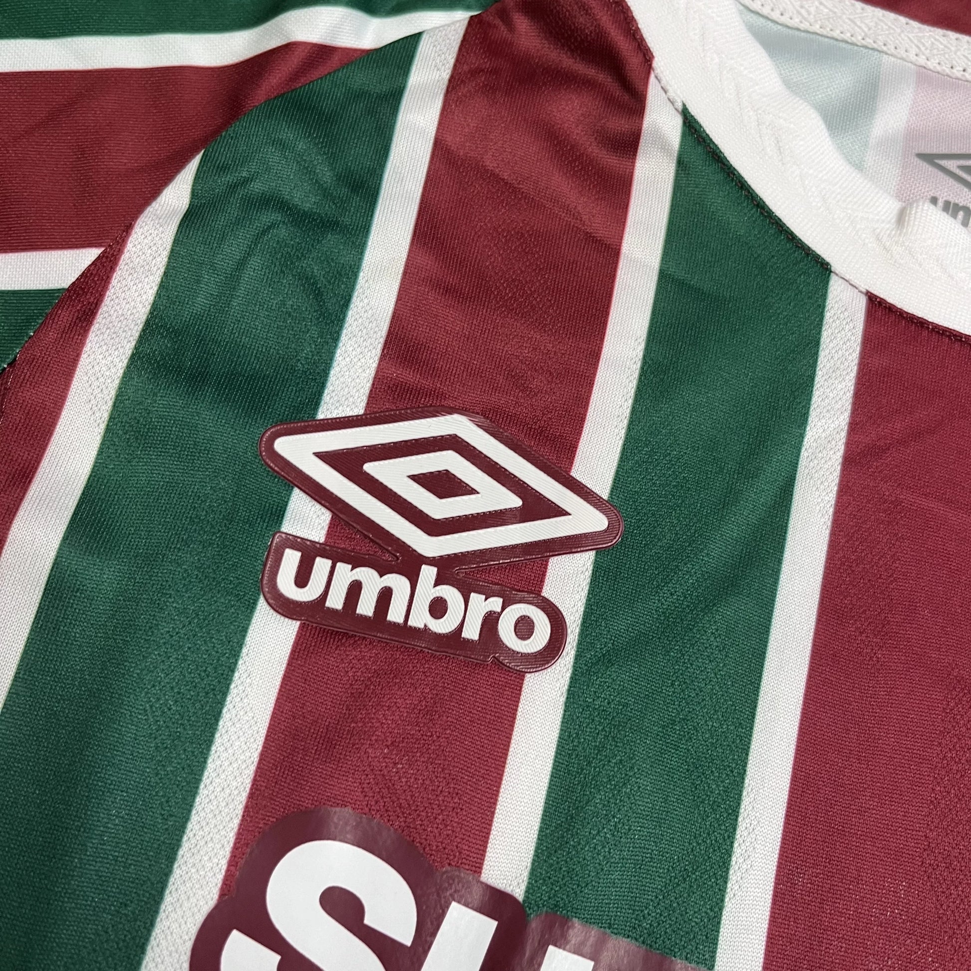 Close do logotipo da Umbro bordado em branco com contorno grená, posicionado sobre uma das listras verdes da camisa.