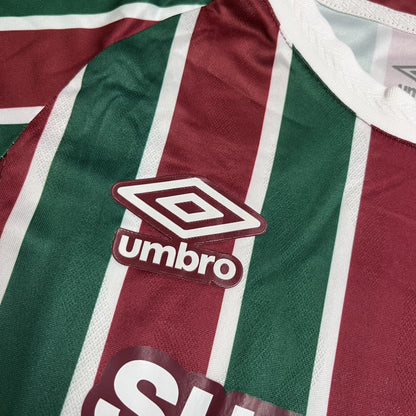 Close do logotipo da Umbro bordado em branco com contorno grená, posicionado sobre uma das listras verdes da camisa.