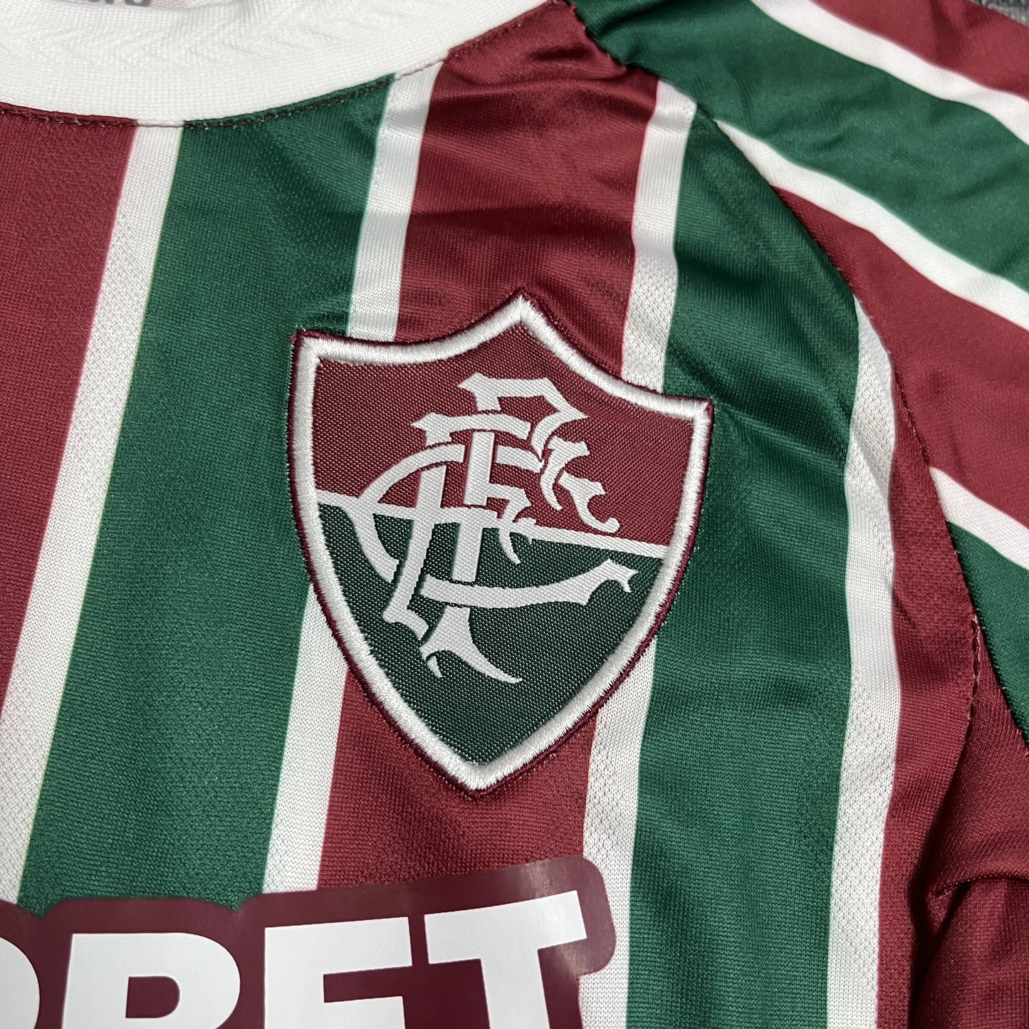 Close detalhado do escudo clássico do Fluminense Football Club aplicado sobre as listras tricolores da camisa.