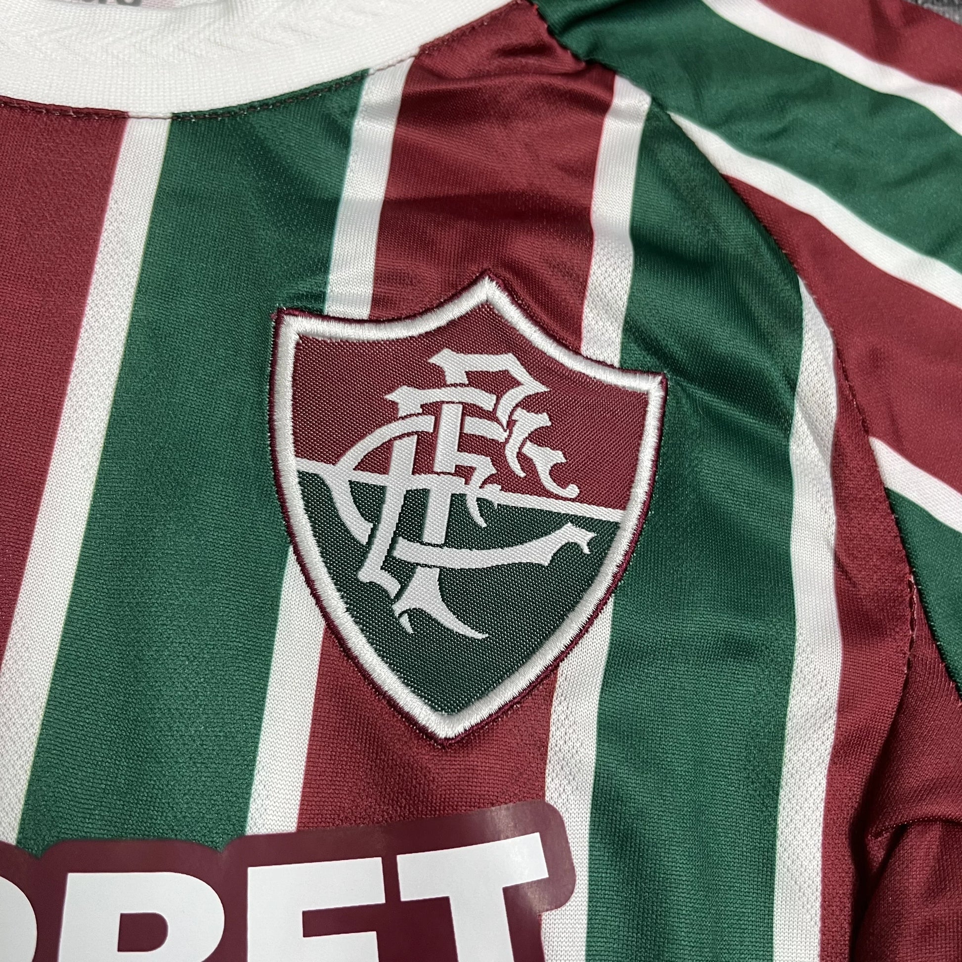 Close detalhado do escudo clássico do Fluminense Football Club aplicado sobre as listras tricolores da camisa.