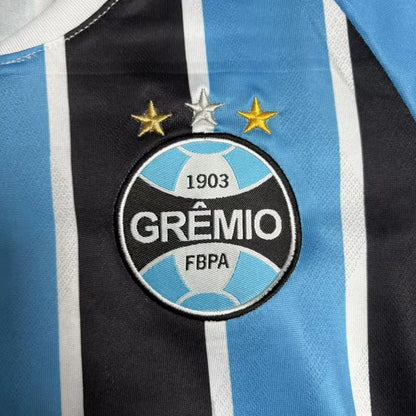Close detalhado do escudo oficial do Grêmio FBPA com suas três estrelas (uma dourada, uma prateada e uma de bronze), aplicado centralizadamente sobre a listra preta da camisa.