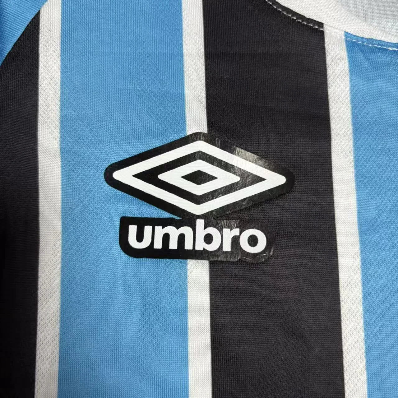 Close do logotipo da Umbro bordado em branco com contorno preto, posicionado sobre uma das listras azuis da camisa.