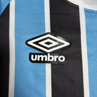 Close do logotipo da Umbro bordado em branco com contorno preto, posicionado sobre uma das listras azuis da camisa.