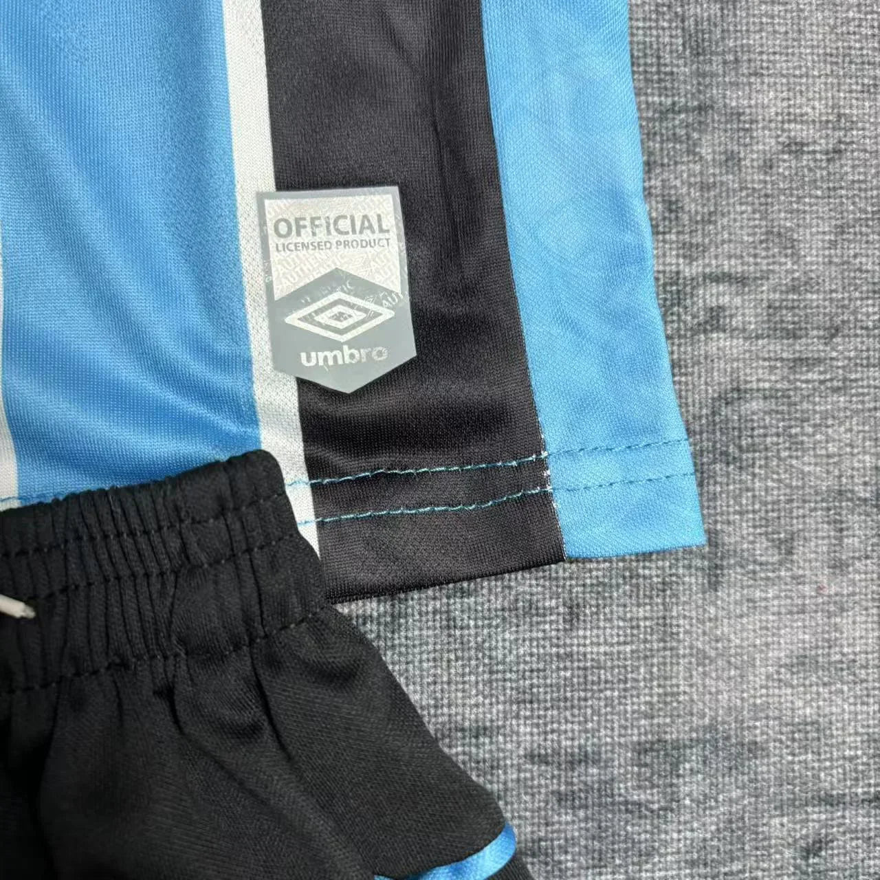 Detalhe lateral da barra da camisa mostrando o selo holográfico de autenticidade da Umbro próximo à costura, acima do calção preto.