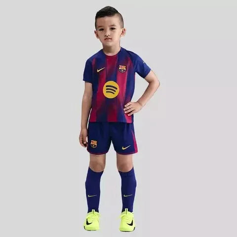 Menino veste kit infantil Barcelona listrado em azul marinho e vermelho, com escudo do time, shorts e meias azuis, chuteiras amarelas.