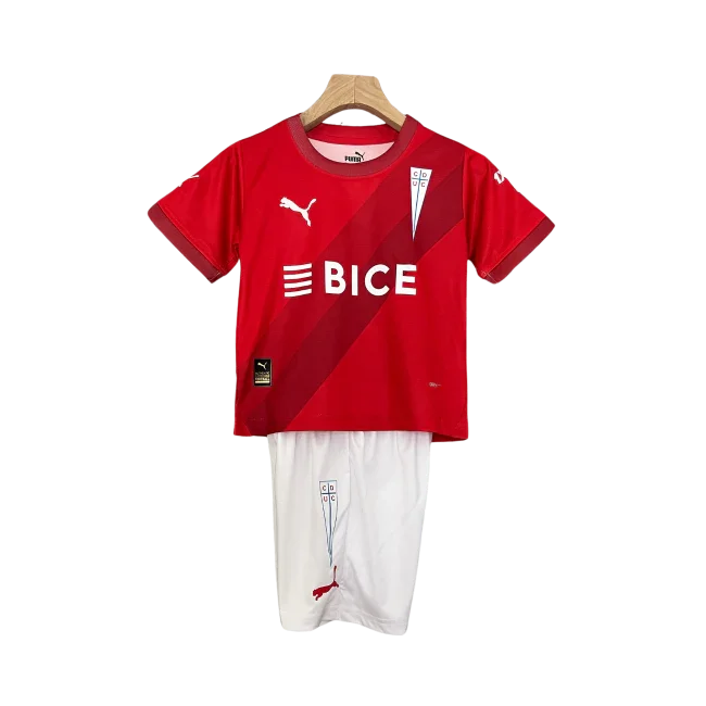 Kit infantil da Universidad Católica 24/25, com camisa vermelha com faixas diagonais, logo BICE e shorts brancos, pendurado.