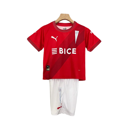 Kit infantil da Universidad Católica 24/25, com camisa vermelha com faixas diagonais, logo BICE e shorts brancos, pendurado.