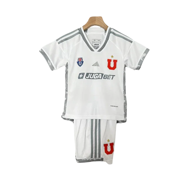 Kit infantil branco da Universidade do Chile, com camisa e shorts. Camisa com listras cinzas nos ombros, logos Adidas, Juga Bet, escudo e 'U' vermelho. Shorts brancos com 'U' vermelho.