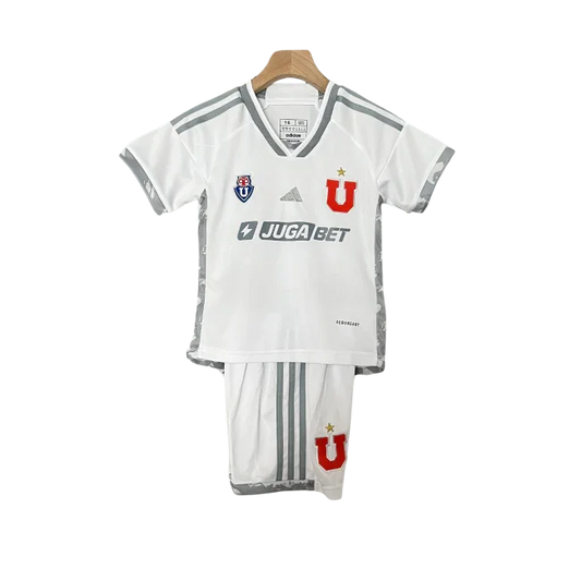 Kit infantil branco da Universidade do Chile, com camisa e shorts. Camisa com listras cinzas nos ombros, logos Adidas, Juga Bet, escudo e 'U' vermelho. Shorts brancos com 'U' vermelho.